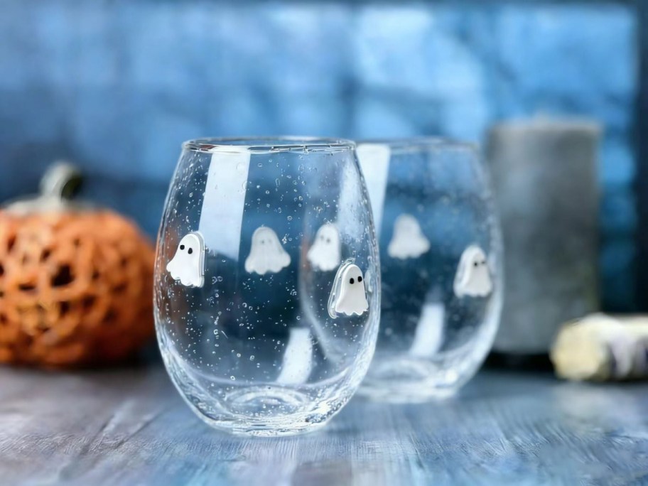 Two clear, bubbly stemless wine glasses decorated with small white ghosts, sitting on a wooden surface with an orange pumpkin in the background.