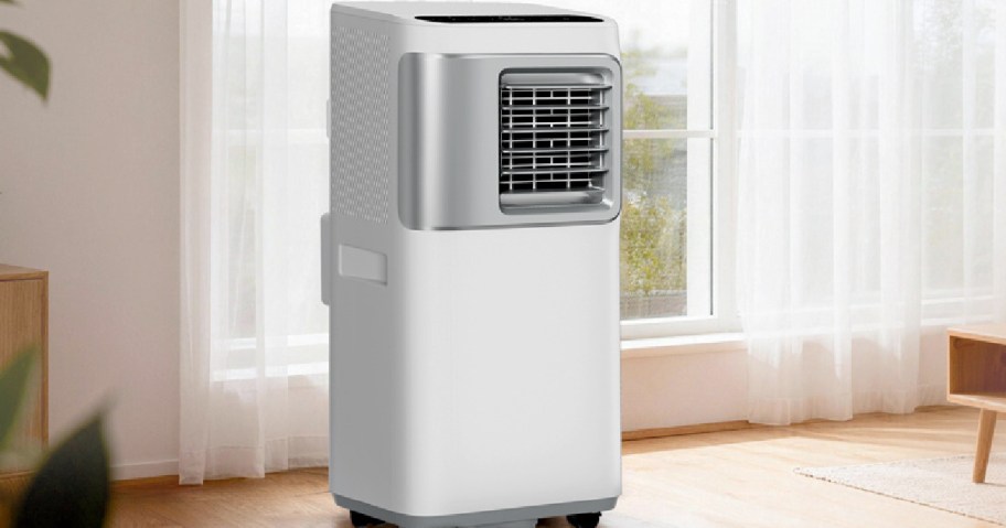 A white portable air conditioner standing on a light wood floor in front of a window with sheer white curtains.