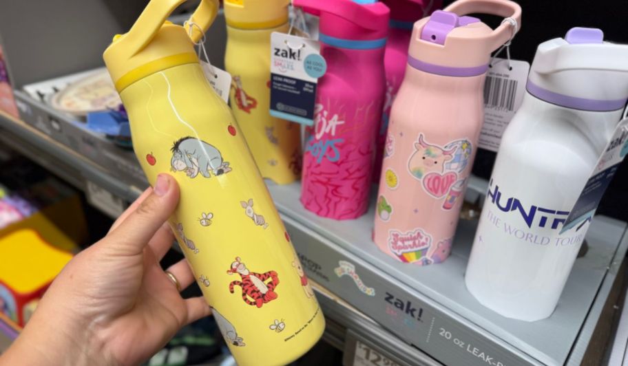 Hand holding Winnie the Pooh Zak! 20oz Character Stainless Steel Bottle next to multiple bottle son display at the store