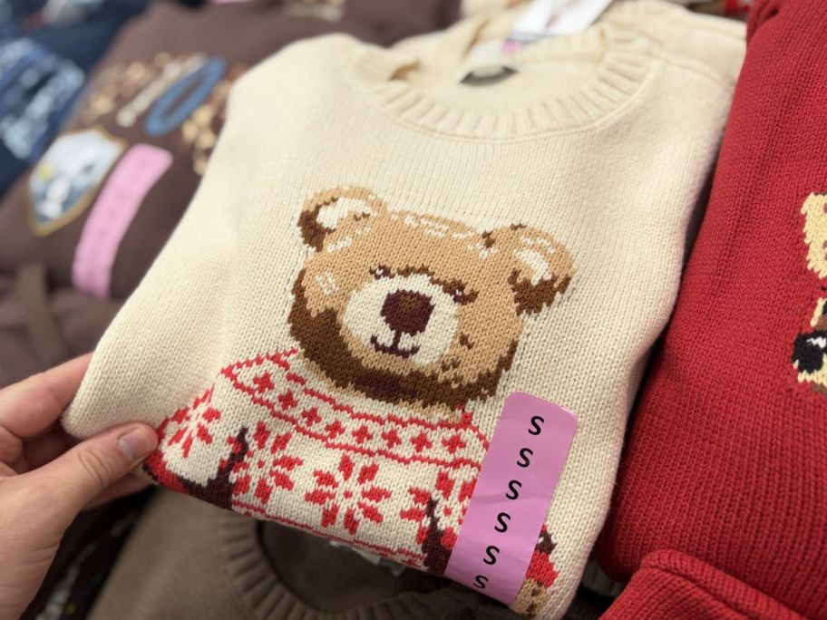 woman holding beige bear sweater next to a red sweater