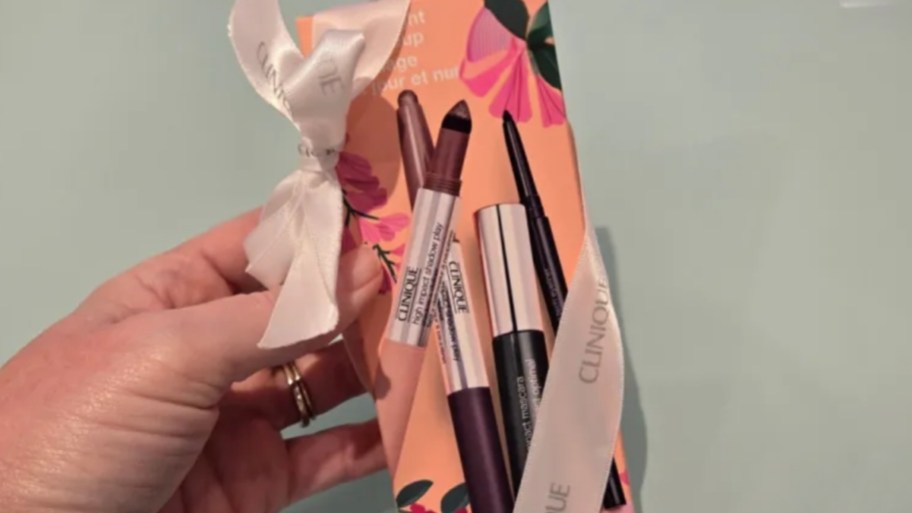 A hand holds a Clinique makeup gift set adorned with a white ribbon. The box features floral designs and showcases three cosmetic products on the cover.