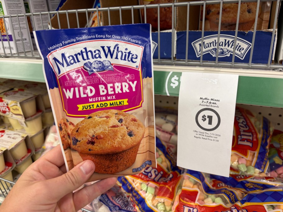 A hand holding a pouch of Martha White Wild Berry muffin mix in a store aisle next to a hanging paper sign advertising a $1 sale price.