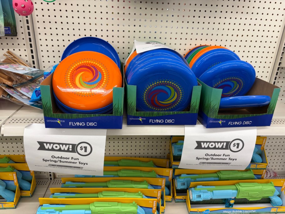 A store shelf displaying colorful plastic flying discs and toy water guns with hanging paper signs advertising a $1 sale on outdoor spring and summer toys.