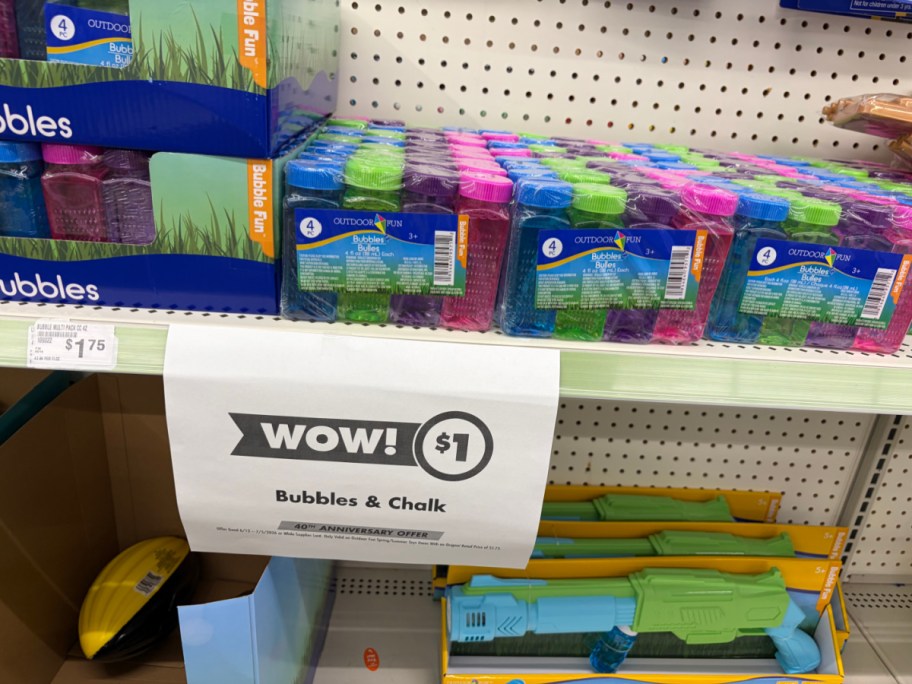A store shelf displaying multi-packs of colorful bubbles and toy water guns with a hanging paper sign advertising a $1 sale on bubbles and chalk.
