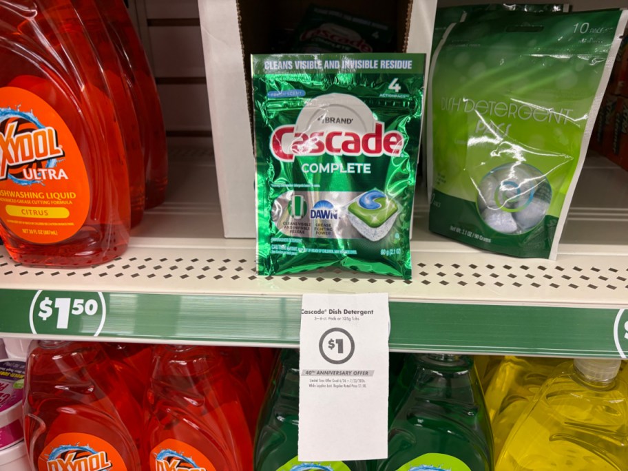 A store shelf displaying a small green pouch of Cascade Complete dishwasher detergent pacs with a hanging paper sign advertising a $1 sale price.