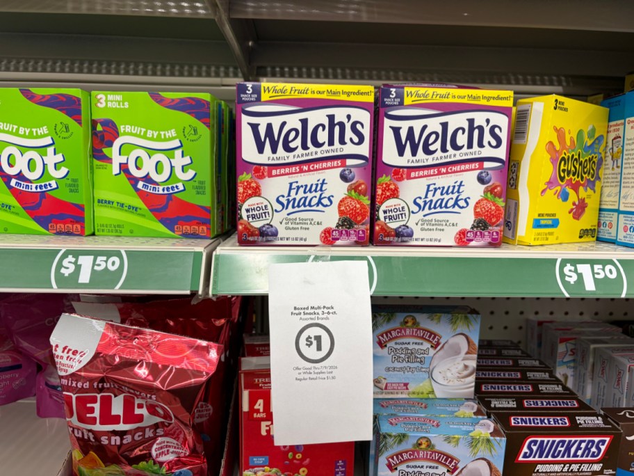 A store shelf displaying boxes of fruit snacks, including Welch's and Fruit by the Foot, with a hanging paper sign advertising a $1 sale price.