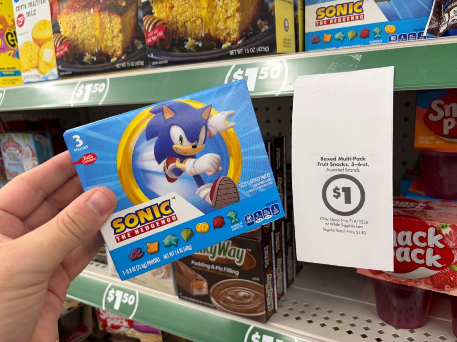 A hand holding a box of Sonic the Hedgehog fruit snacks in a store aisle next to a hanging paper sign advertising a $1 sale price.