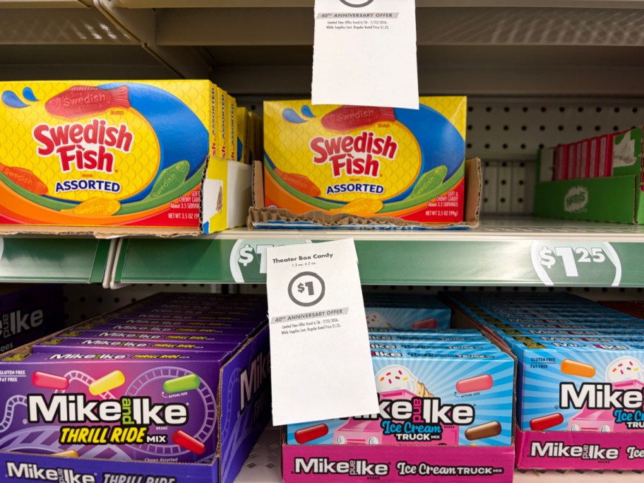 A store shelf displaying theater boxes of Swedish Fish and Mike and Ike candies with a hanging paper sign advertising a $1 sale price.
