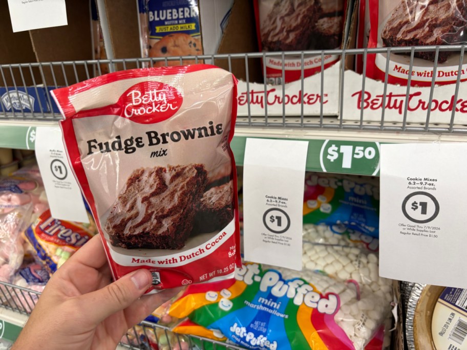 A hand holding a pouch of Betty Crocker Fudge Brownie mix in a store aisle next to hanging paper signs advertising a $1 sale price.