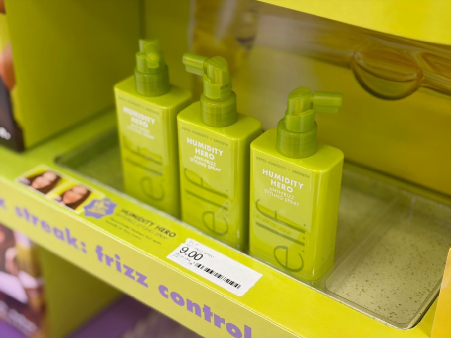 Three bright green bottles of e.l.f. Humidity Hero Anti-Frizz Styling Spray lined up on a yellow store display shelf.