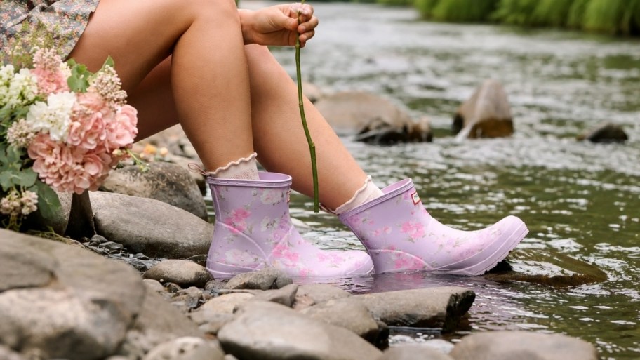 a women sitting on the side of a stream in a pair of short rain boots