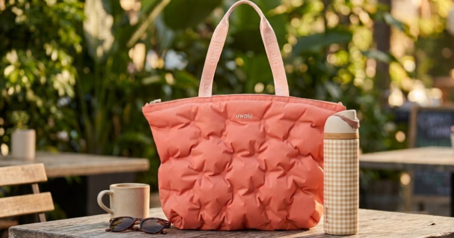 A pink puffy quilted Owala tote bag next to a tan gingham Owala water bottle, a beige mug, and tortoiseshell sunglasses resting on an outdoor wooden table.