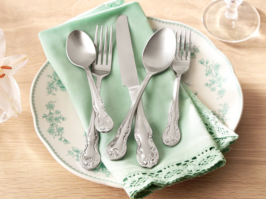 A five-piece set of silver flatware resting on a light green napkin over a white plate with green floral designs.