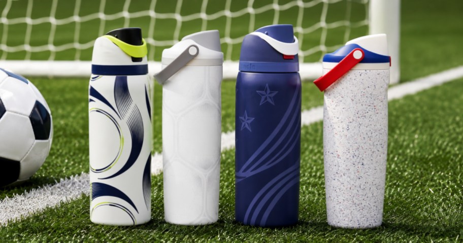 Four Owala water bottles with soccer-themed designs standing on a grassy soccer field next to a soccer ball, with a white field line and goal net in the background.