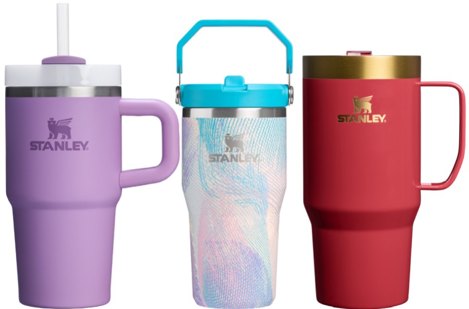 Three Stanley tumblers side-by-side: a light purple tumbler with a straw and side handle, a pastel patterned tumbler with a bright blue lid and carry handle, and a solid red tumbler with a gold rim and side handle.
