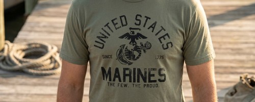 A person wearing an olive green United States Marines t-shirt standing on a wooden dock.