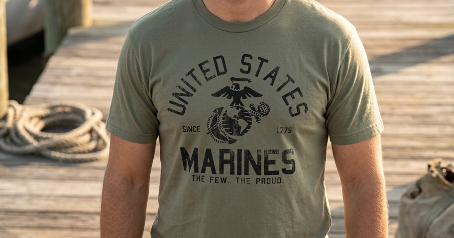 A person wearing an olive green United States Marines t-shirt standing on a wooden dock.