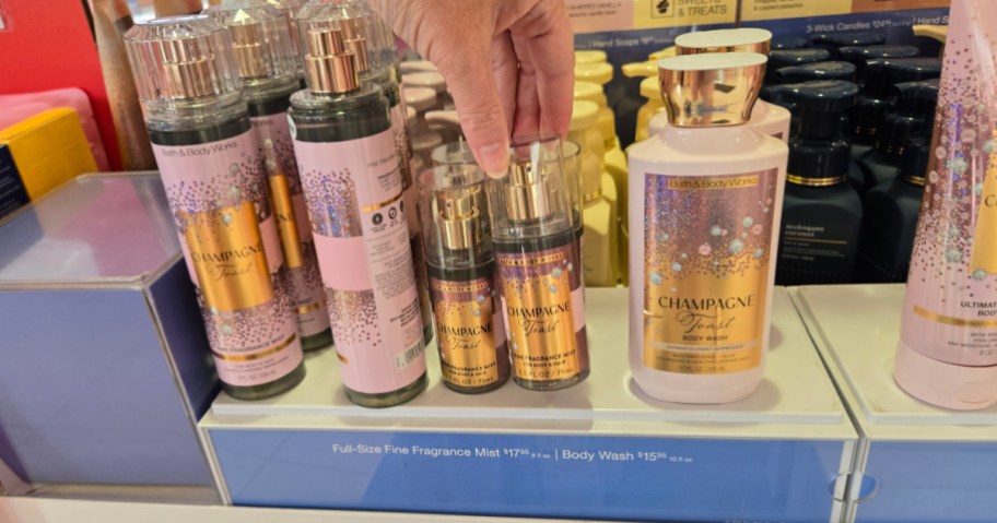 A hand picking up a mini bottle of Bath & Body Works Champagne Toast fragrance mist from a store display surrounded by other matching products.