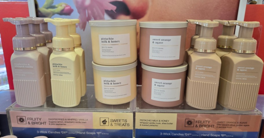 A store display shelf featuring bottles of White Barn foaming hand soap alongside stacks of matching 3-wick candles in various pastel colors and scents.