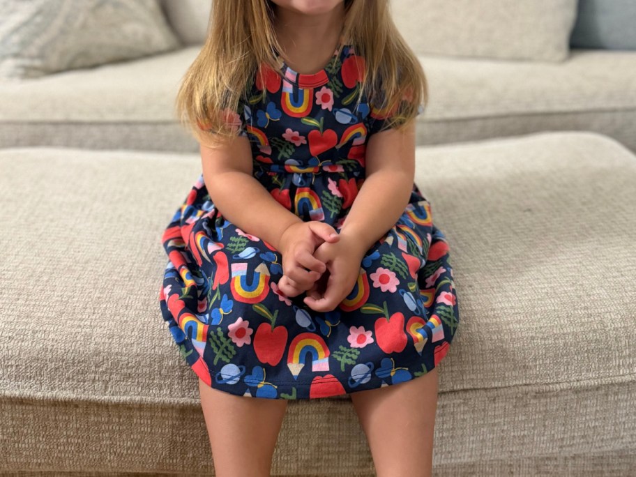 A young child sitting with hands clasped, wearing a dark blue short-sleeved dress with a colorful pattern of rainbows, apples, and flowers.