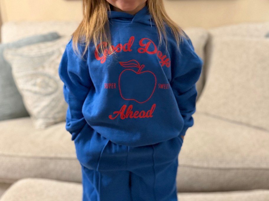 A child standing in front of a couch wearing a blue hoodie and matching sweatpants. The hoodie features a red apple graphic and the words "Good Days Ahead" in red text.