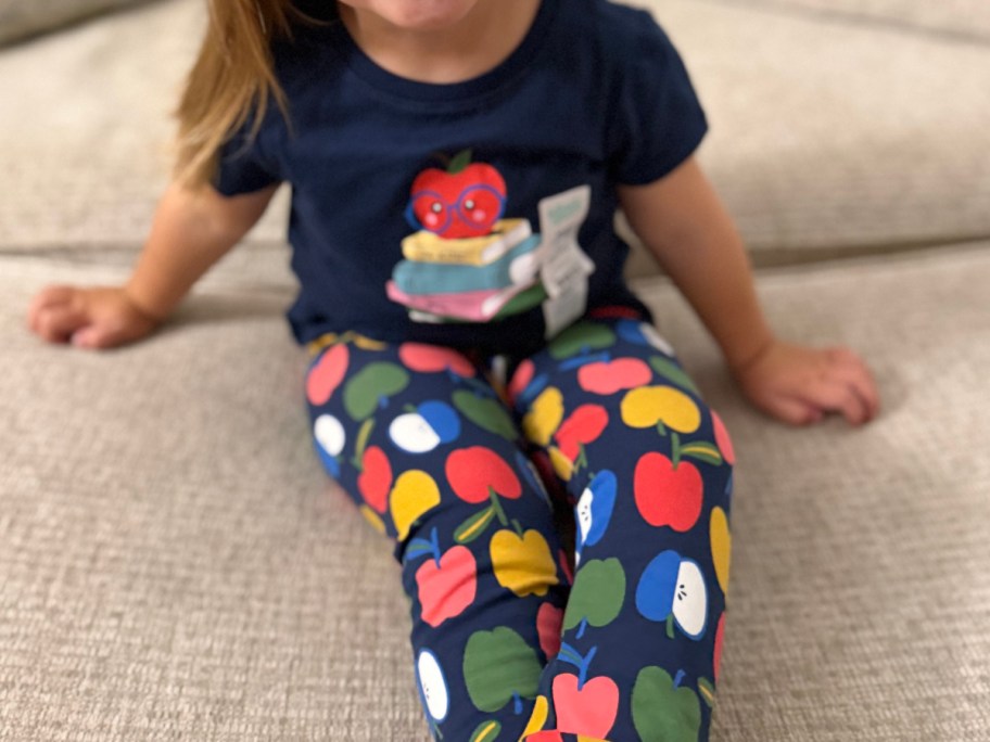 A young child sitting on a couch wearing a navy blue t-shirt with an apple and books graphic, paired with navy leggings featuring a colorful apple pattern.