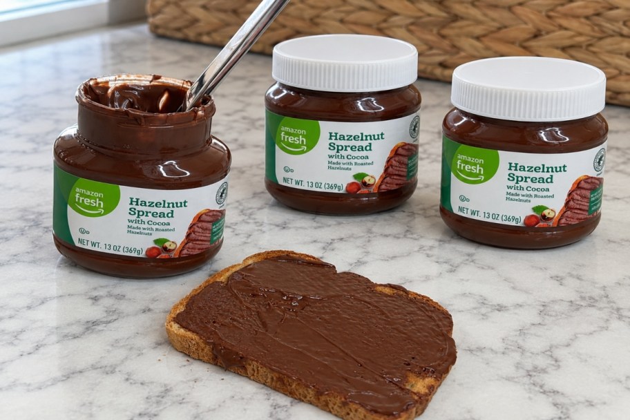 Three Jars of Amazon Fresh Hazelnut Spread on the counter. One open with a butter knife in it. One piece of bread with the spread on it.
