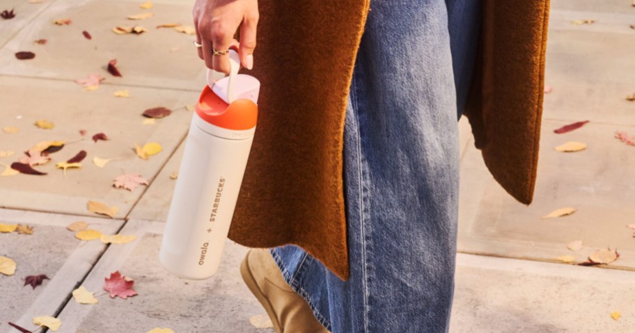 A person walking on a sidewalk strewn with autumn leaves, holding a cream-colored Owala x Starbucks water bottle with an orange lid.