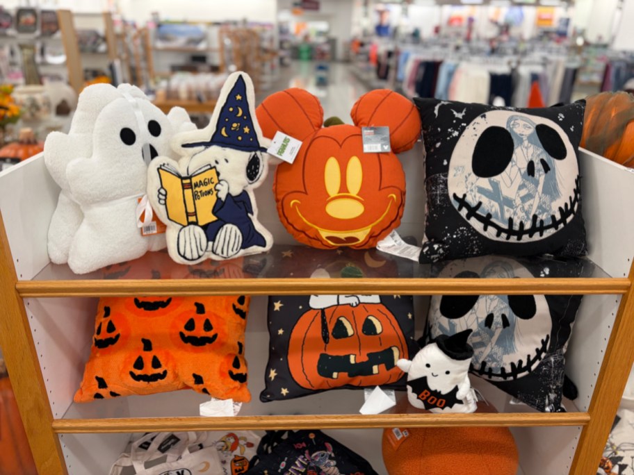 A store display shelf filled with various Halloween-themed decorative pillows, including a white ghost, Snoopy reading a spellbook, a Mickey Mouse pumpkin shape, and a Jack Skellington pillow.