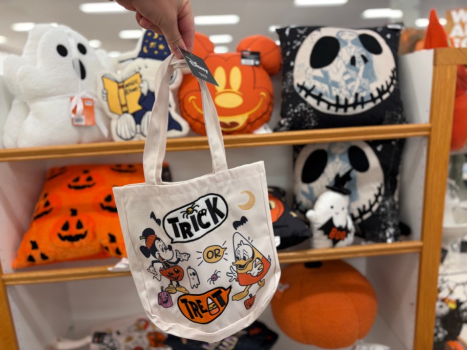 A hand holding a white canvas Disney Halloween trick-or-treat tote bag featuring illustrations of Mickey Mouse and Donald Duck with festive icons and "TRICK OR TREAT" text.
