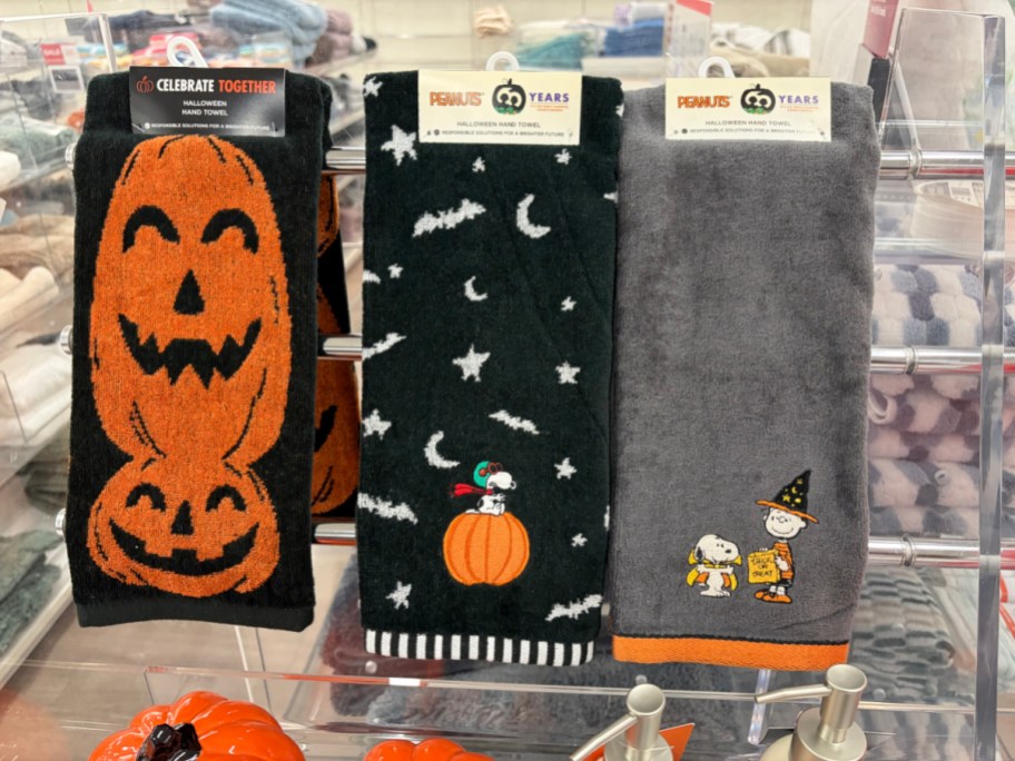 Three Peanuts and Celebrate Together Halloween hand towels hanging on display, featuring a black towel with stacked jack-o'-lanterns, a black towel with bats, stars, and Snoopy on a pumpkin, and a grey towel with Charlie Brown in a witch hat and Snoopy in a cape holding a trick-or-treat bucket.