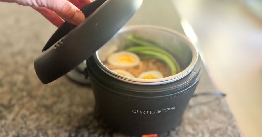 Hand opening the lid on a Curtis Stone Food warmer with ramen, eggs, and green onions inside.