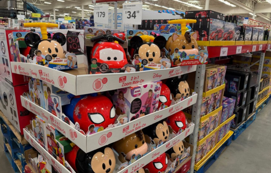 A store display shelf stocked with various Disney and character 2-in-1 Zoom Ride-On toys, including Mickey Mouse, Cars, Spider-Man, and Bullseye styles.