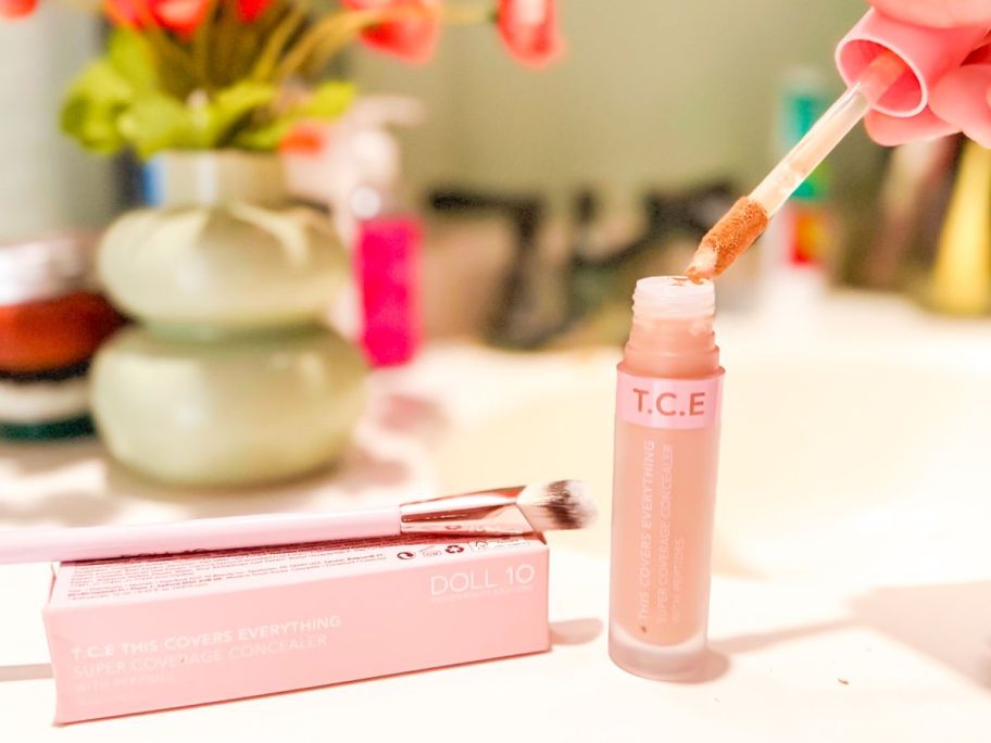 hand removing applicator from Doll 10 T.C.E. Super Coverage Concealer with brush on top of box next to it