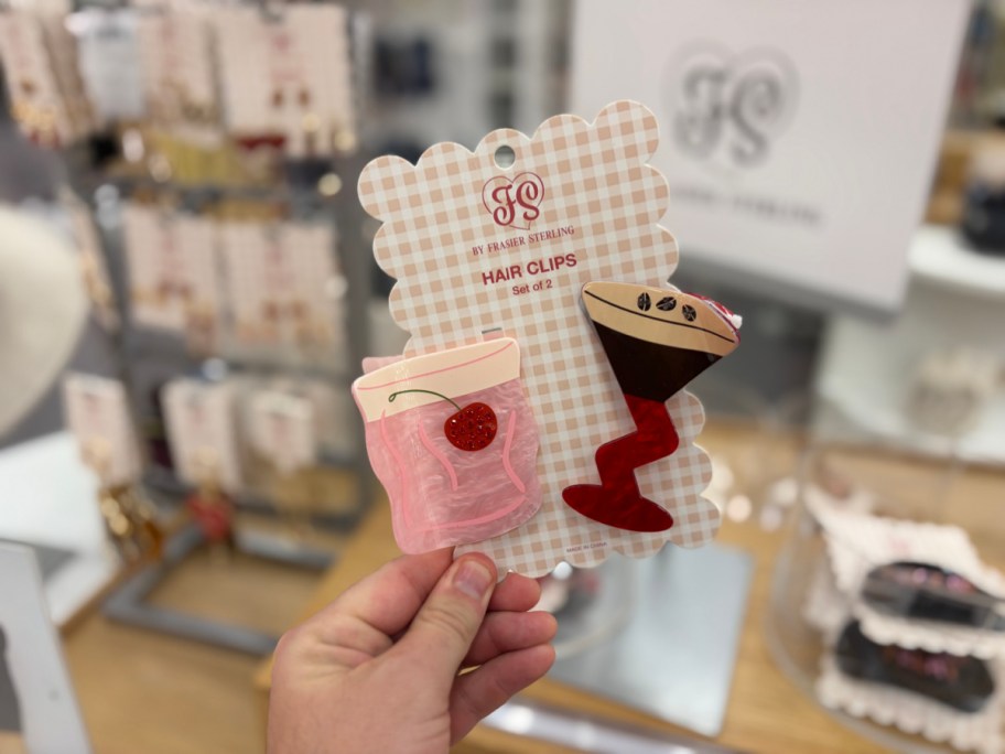 A hand holding a two-piece set of cocktail-themed hair claw clips featuring a pink drink with a cherry and an espresso martini on a gingham backing card.