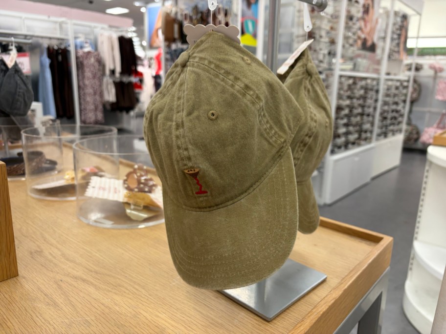 An olive green baseball hat with a small embroidered espresso martini graphic on the front, displayed on a stand in a retail store.
