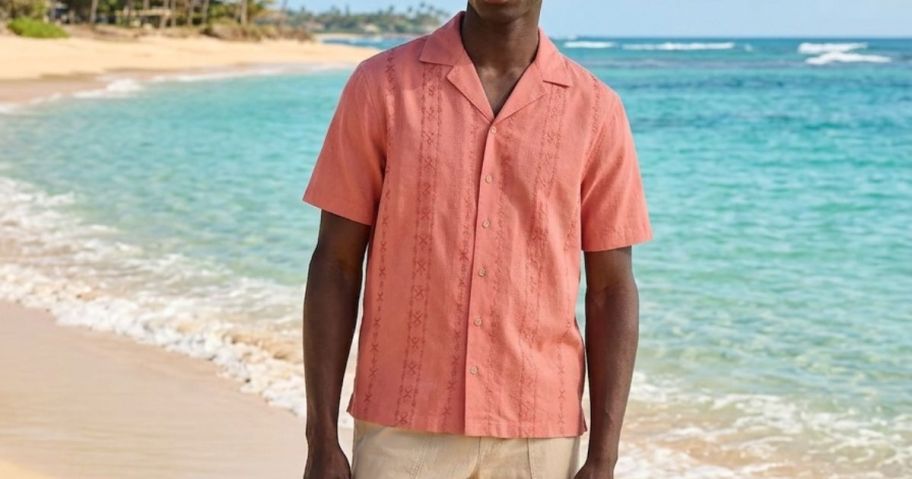 man wearing soft salmon colored George Men's Embroidered Camp Shirt on beach
