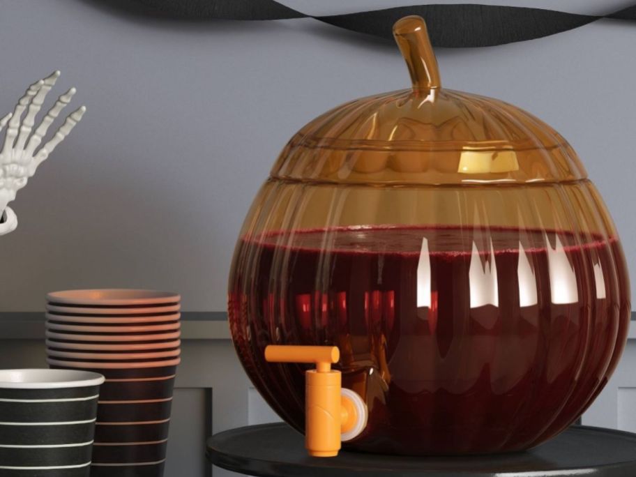 Halloween pumpkin dispenser displaye don top of a table next to cups and a skeleton hand