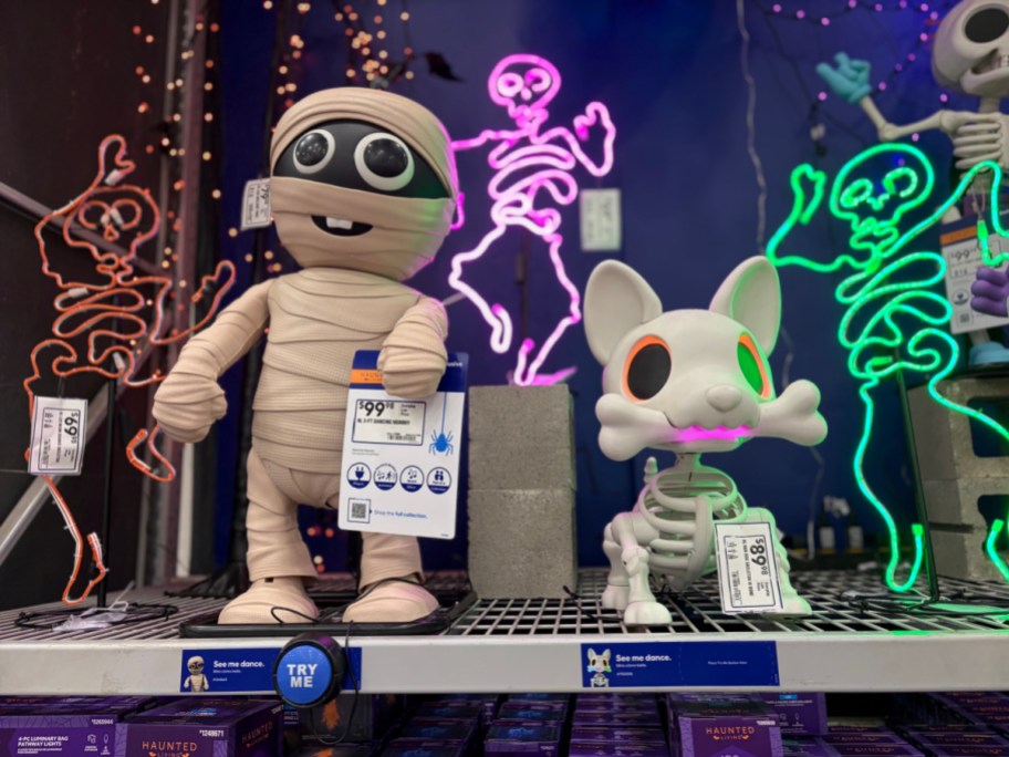 A mummy animatronic holding a price tag next to a skeleton dog animatronic on a store shelf decorated with neon Halloween lights.