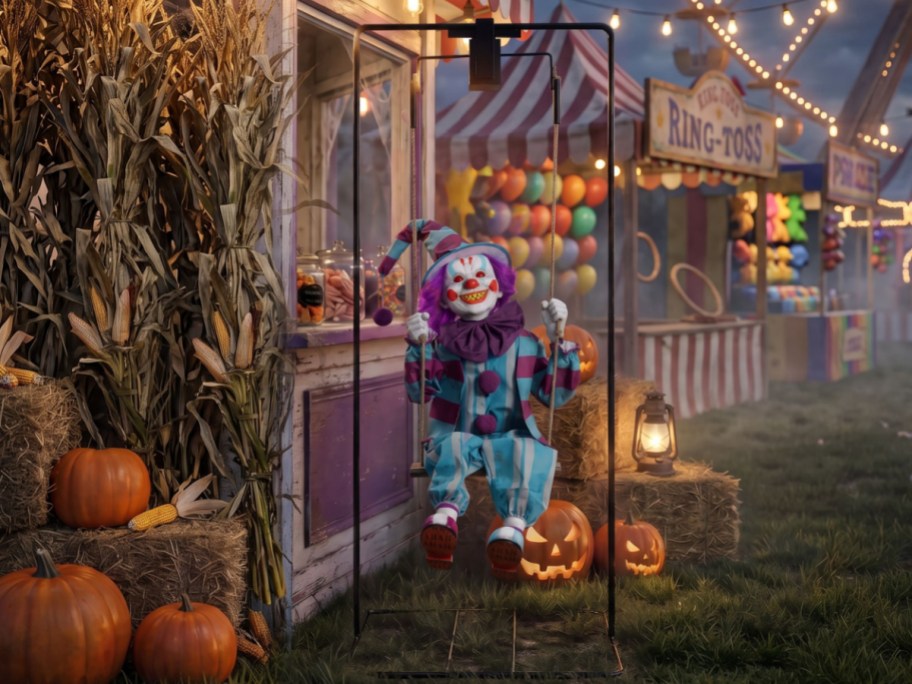 A scary clown animatronic in a blue and purple striped outfit on a swing set at a carnival decorated with pumpkins, corn stalks, and string lights.