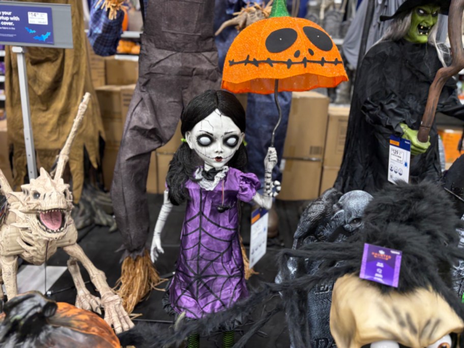 A creepy doll animatronic wearing a purple spiderweb dress and holding a bright orange pumpkin-shaped umbrella in a store display.