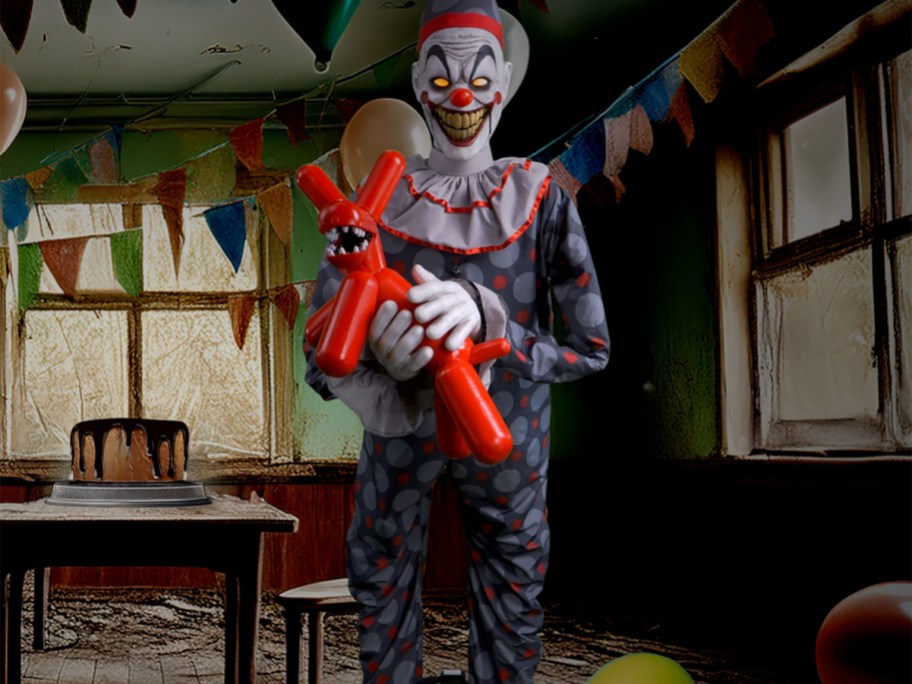 A creepy clown animatronic wearing a polka-dot outfit and holding a red balloon animal dog in a party room decorated with party banners and a cake on a table.