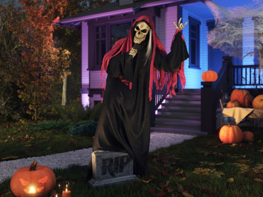 A grim reaper skeleton animatronic wearing a black and red robe, holding a microphone while standing on a tombstone in a yard decorated with pumpkins.