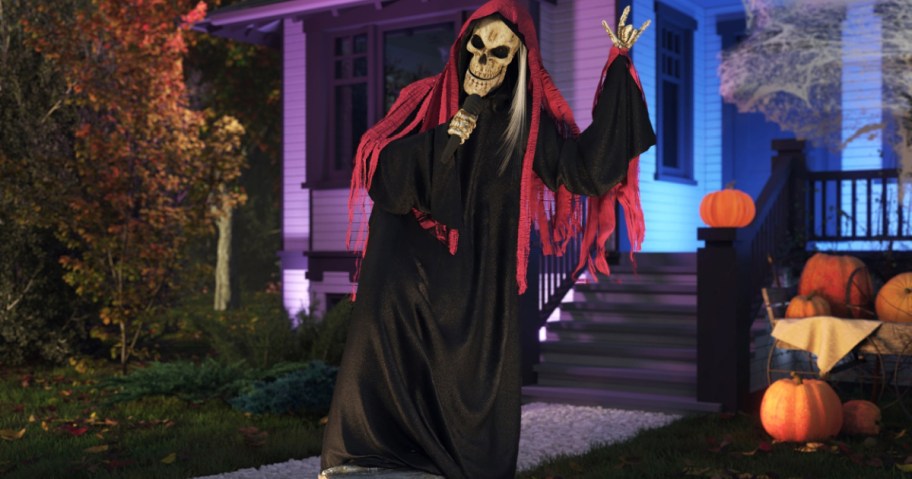 A 7-foot light-up reaper band singer animatronic wearing a black robe and red hood while holding a microphone outside a house decorated with pumpkins.