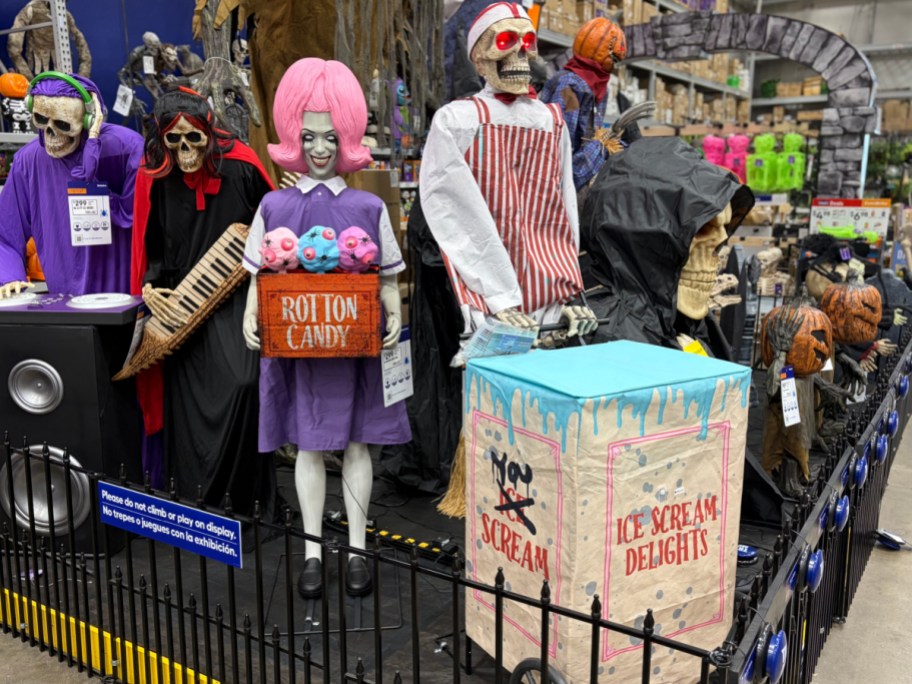 Halloween animatronics displayed in a store, including a girl holding a box labeled "Rotton Candy," a skeleton in an apron, a hooded figure, and an ice cream cart labeled "Ice Scream Delights."