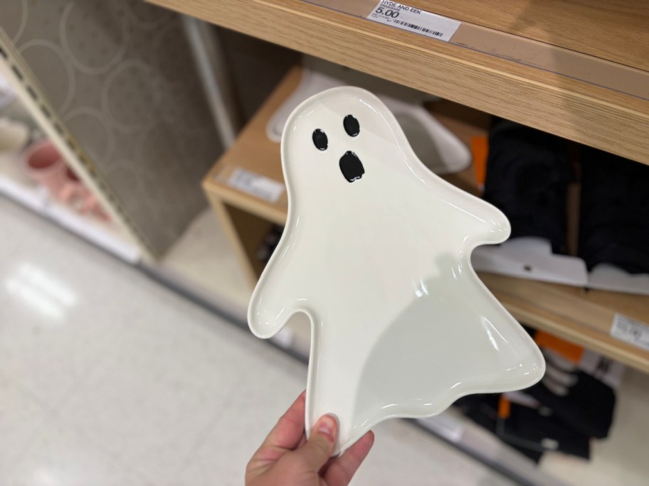 A hand holding a white ghost-shaped Hyde & EEK! Halloween stoneware serving platter with a spooky face.