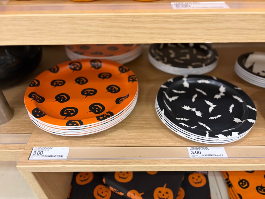 Stacks of orange Hyde & EEK! Halloween melamine dinner plates with black pumpkin patterns and black plates with white bat patterns displayed on store shelves.