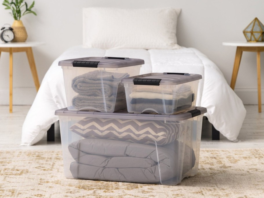 Clear plastic storage boxes with gray lids filled with folded linens and bedding, stacked on a rug in a bedroom.