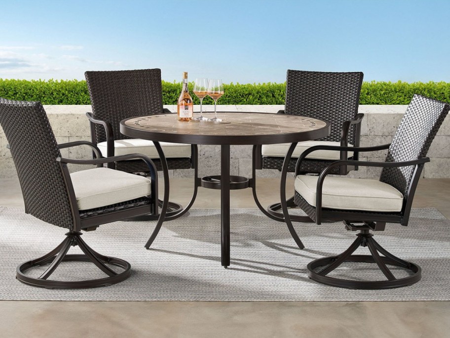 An outdoor dining set with a round table, a bottle of wine, two wine glasses, and four wicker swivel chairs with white cushions on a patio.