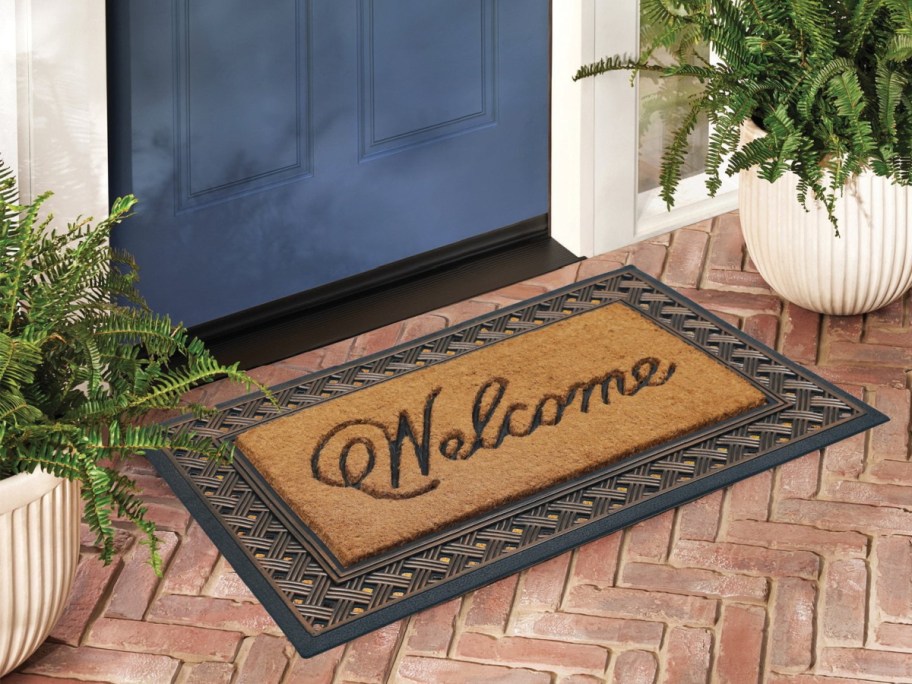 A coir doormat with the word "Welcome" in cursive, surrounded by a decorative woven frame, sitting outside a blue front door on a brick porch.